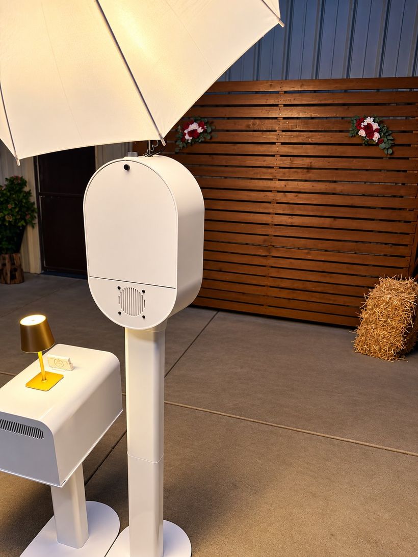 Photo booth with umbrella light and print station set up outdoors at dusk beside a wood slat backdrop with florals